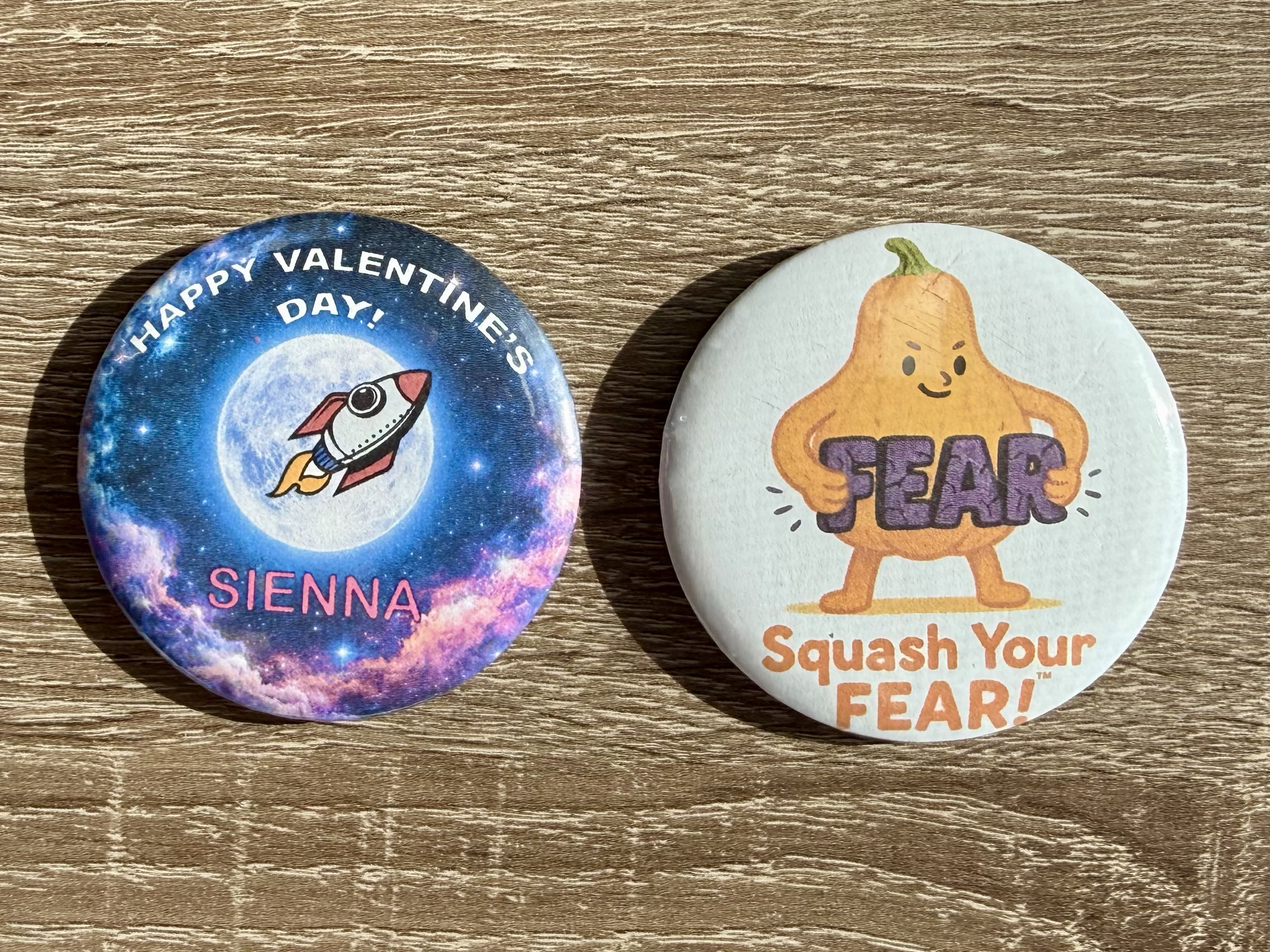 Two finished circular buttons on a wooden surface: a space-themed Valentine design and a Squash Your Fear design.